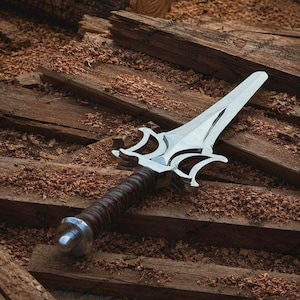 May include: A silver-colored metal dagger featuring a pointed blade with a complex, symmetrical guard design. The handle is wrapped in dark brown material and finished with a rounded silver-colored pommel at the base of the grip.