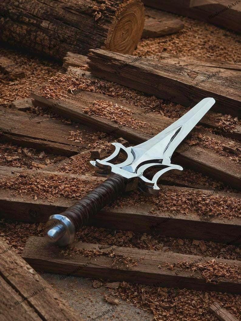 May include: A silver-colored metal dagger featuring a pointed blade with a complex, symmetrical guard design. The handle is wrapped in dark brown material and finished with a rounded silver-colored pommel at the base of the grip.