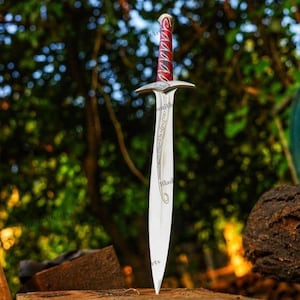 May include: A silver-toned metal sword featuring a pointed blade with intricate etched scrollwork patterns. The hilt includes a crossguard and a handle wrapped in a vibrant red material with silver accents, culminating in a rounded pommel.