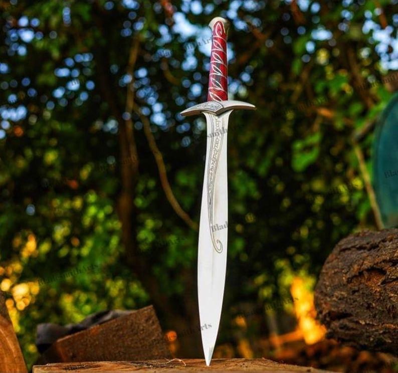 May include: A silver-toned metal sword featuring a pointed blade with intricate etched scrollwork patterns. The hilt includes a crossguard and a handle wrapped in a vibrant red material with silver accents, culminating in a rounded pommel.