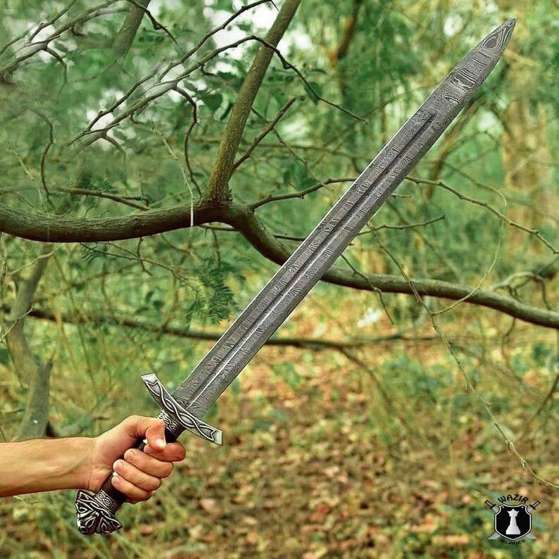 May include: A silver-colored sword with a detailed blade and handle is held in a hand. The sword has a textured pattern on the blade and ornate designs on the handle. The background features a blurred view of trees and foliage.