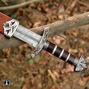 May include: A detailed close-up of a Viking sword with a patterned blade and ornate silver accents. The handle is dark brown with silver bands. The sword rests on a tree branch, showcasing its craftsmanship.