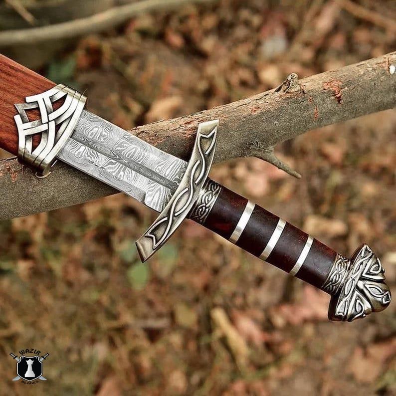 May include: A detailed close-up of a Viking sword with a patterned blade and ornate silver accents. The handle is dark brown with silver bands. The sword rests on a tree branch, showcasing its craftsmanship.