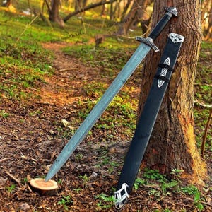May include: A long, silver sword with a black handle and a matching black sheath rests against a tree. The sword has intricate silver detailing on the handle and crossguard. The sheath has silver accents and a decorative design.