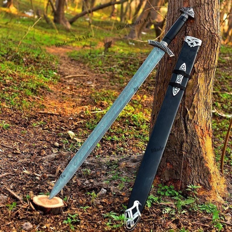 May include: A long, silver sword with a black handle and a matching black sheath rests against a tree. The sword has intricate silver detailing on the handle and crossguard. The sheath has silver accents and a decorative design.