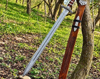 May include: A double-edged steel sword with a patterned blade, a crossguard, and a brown leather-wrapped handle. Beside it stands a matching wooden scabbard featuring intricate carved knotwork patterns and silver-toned metal accents.