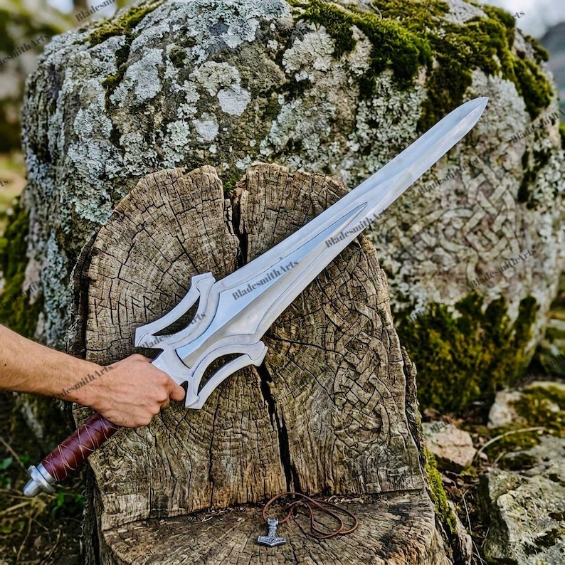 May include: A silver-colored metal sword with a pointed blade and a complex, open-work crossguard. The handle is wrapped in brown leather. The blade features the engraved text BladesmithArts. A small metal hammer pendant rests below the sword.