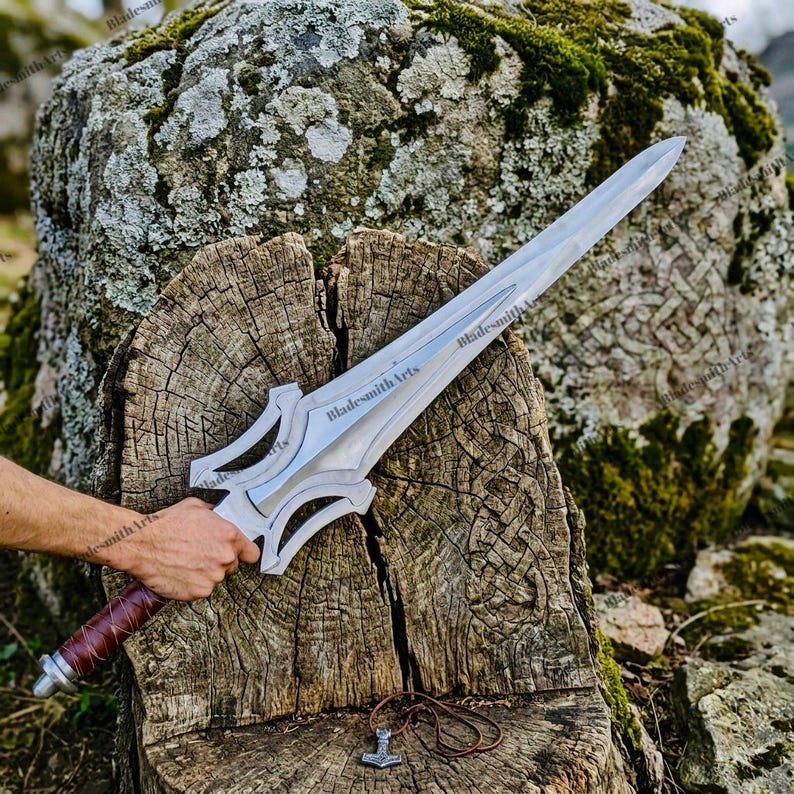 May include: A silver-colored metal sword with a pointed blade and a complex, open-work crossguard. The handle is wrapped in brown leather. The blade features the engraved text BladesmithArts. A small metal hammer pendant rests below the sword.