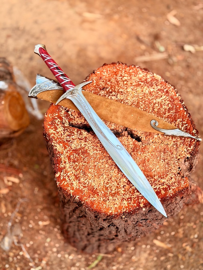 May include: A silver-toned sword with a pointed blade and a crossguard featuring intricate engravings. The handle is deep red with silver accents. A tan-colored scabbard with silver metal detailing at the tip and throat rests beneath the sword.