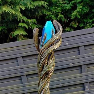 Op de afbeelding: Een houten staf met een gedraaid ontwerp, met een helderblauwe kristal aan de bovenkant. De staf is bruin met groene accenten aan de basis, tegen een donkere houten schutting en groen gebladerte.