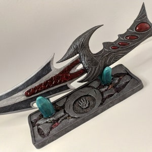 May include: A silver and red fantasy dagger with a black and red handle, displayed on a grey stone base with two teal gems.