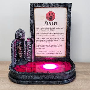 May include: A 3D printed model of a dark brotherhood shrine with a red light illuminating the base. The shrine features a stone archway with a figure inside and a plaque with the text "Tenets" and the five tenets of the Dark Brotherhood.