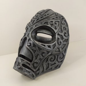 Puede incluir: Una máscara gris con intrincados patrones arremolinados y una abertura ovalada para los ojos. La máscara tiene una apariencia ligeramente desgastada, lo que sugiere un tema histórico o de fantasía. El diseño es simétrico y detallado.