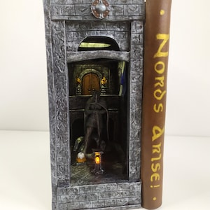 May include: A miniature book with a stone castle facade and a glowing interior. The book spine reads "Nords Arise!" in gold lettering on a brown background.
