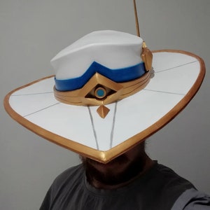 May include: A white hat with a wide brim and a gold trim. The hat has a blue band and a gold and blue design on the front. The hat is worn by a person with a beard.