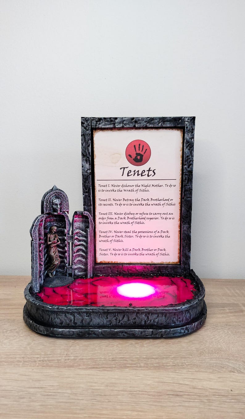May include: A 3D printed model of a dark brotherhood shrine with a red light illuminating the base. The shrine features a stone archway with a figure inside and a plaque with the text "Tenets" and the five tenets of the Dark Brotherhood.