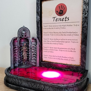 May include: A dark grey stone fountain with a red liquid inside. The fountain has a small statue of a woman inside. A framed piece of paper with the text "Tenets" and a list of rules is behind the fountain.
