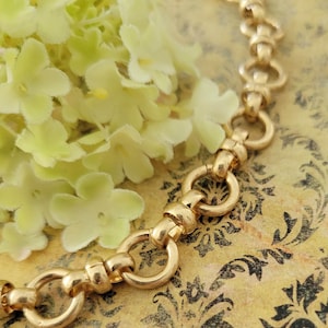 May include: A gold-toned necklace with a series of interlocking circular and oblong links. The necklace is displayed on a patterned background with soft yellow flowers in the upper left corner. The necklace has a polished finish.