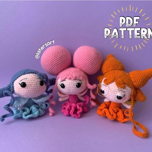 Peut inclure: Trois poupées amigurumi crochetées avec des cheveux et des tenues de différentes couleurs. La poupée à gauche est bleue et porte une tenue bleu et blanc. La poupée au milieu est rose et porte une tenue rose et blanc. La poupée à droite est orange et porte une tenue orange et blanc. Les poupées sourient toutes et ont de grands yeux. Le texte "PDF PATTERN" est dans le coin supérieur droit de l'image.