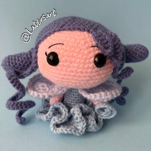 May include: A crocheted doll with a round light pink head, black plastic eyes, and purple yarn hair styled in side curls. The doll features light blue crocheted wings and a ruffled light blue skirt. The text @latersart is visible in the upper left.