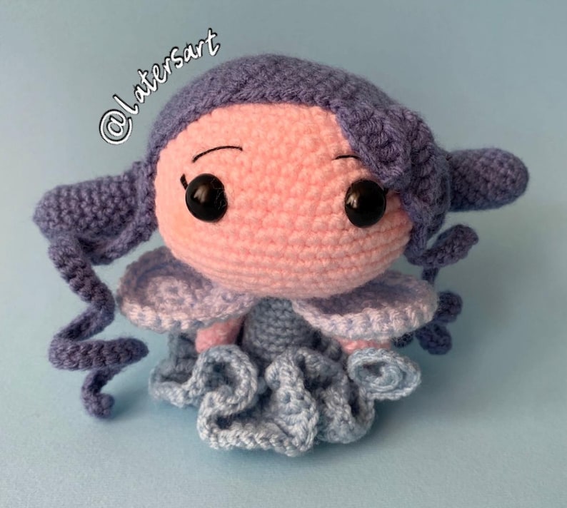 May include: A crocheted doll with a round light pink head, black plastic eyes, and purple yarn hair styled in side curls. The doll features light blue crocheted wings and a ruffled light blue skirt. The text @latersart is visible in the upper left.