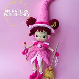 May include: A crochet doll featuring a pink pointed hat, dark brown hair, and a pink dress with a multi-colored circular emblem. The doll holds a multi-colored beaded wand and stands next to a small broom. Text reads: PDF PATTERN ENGLISH ONLY.