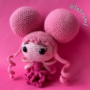 May include: A crochet doll with light pink skin, large black eyes, and two oversized spherical pink buns. The doll features long, curly pink side tendrils, a dark pink dress with a ruffled skirt, and the text @latersart in the upper right corner.