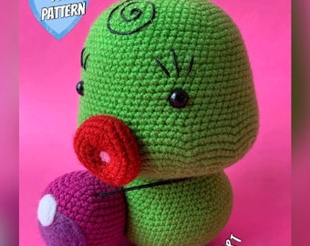 May include: A green crocheted amigurumi figure with a large rounded head, a red circular mouth, and black eyes. It holds a small purple and pink sphere with a white dot. A blue heart icon in the top left corner contains the text PDF PATTERN.