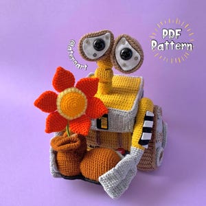 May include: Crochet pattern for a yellow and gray robot holding a red flower. The pattern is available as a PDF download. The robot is holding a flower in a brown pot.