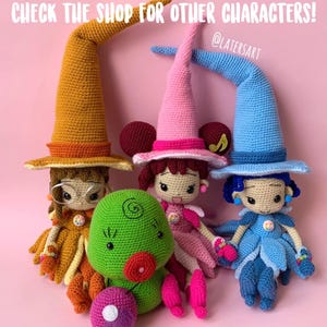 May include: Three crocheted dolls wearing tall pointed hats in mustard yellow, pink, and blue, alongside a round green creature with a red mouth and a purple ball. The text reads: CHECK THE SHOP FOR OTHER CHARACTERS! @LATERSART. Each doll has detailed features.