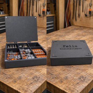 Könnte beinhalten: Eine dunkelgraue Aufbewahrungsbox, geöffnet, um Fächer mit kleinen Hardware-Teilen zu zeigen. Auf dem geschlossenen Deckel steht der Name "Felix" und "Kleinteilebox". Die Box steht auf einer Holzoberfläche.