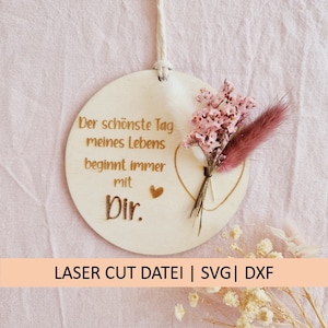 May include: A wooden circle with a heart shape and the text "Der schönste Tag meines Lebens beginnt immer mit Dir."  The circle is decorated with a bouquet of dried pink flowers and a single pink feather.