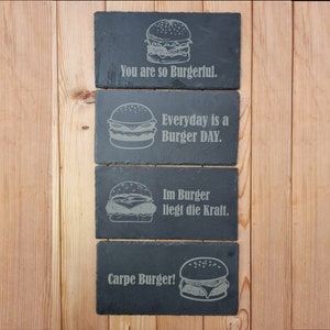 Könnte beinhalten: Vier rechteckige schwarze Schiefer-Servierbretter mit gravierten Burger-Designs und den Sätzen: "You are so Burgerful", "Everyday is a Burger DAY", "Im Burger liegt die Kraft" und "Carpe Burger!". Auf einer Holzoberfläche angeordnet.