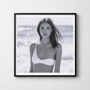 Könnte beinhalten: Schwarz-weiß gerahmtes Foto einer Person, die ein weißes One-Shoulder-Bikini-Oberteil trägt. Die Person steht vor einem verschwommenen Meereshintergrund. Das Bild ist ein Porträt, wobei die Person direkt in die Kamera blickt.