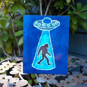 May include: A blue and white watercolor painting of a Bigfoot walking under a UFO. The UFO is a light blue color with a white outline and has a dome on top. The Bigfoot is a dark silhouette with a white outline.