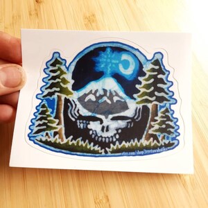May include: A blue and white sticker featuring a mountain scene with a crescent moon. The mountain is surrounded by two pine trees. The sticker has a blue border and the text "etsy.com/shop/treetownbatiks" at the bottom.