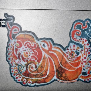 May include: A colorful sticker of an octopus with a batik-style design in shades of orange, red, and white. The octopus is curled up with its tentacles intertwined.