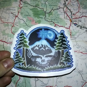 May include: A sticker featuring a blue and white mountain scene with a crescent moon and stars. The mountain is surrounded by two green trees and a dark blue outline. The text "etsy.com/shop/treetownbatiks" is printed at the bottom of the sticker.
