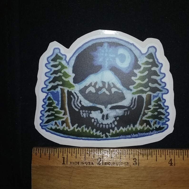 May include: A sticker featuring a blue and green watercolour illustration of a skull with a mountain range and a crescent moon in the background. The skull is surrounded by two pine trees. The text "etsy.com/shop/treetownbahiks" is at the bottom of the sticker.