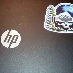May include: A black laptop with a white HP logo on the lid. A sticker of a blue and green mountain scene with a skull and the text "stay calm and follow the music" is on the right side of the lid.