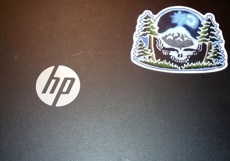 May include: A black laptop with a white HP logo on the lid. A sticker of a blue and green mountain scene with a skull and the text "stay calm and follow the music" is on the right side of the lid.
