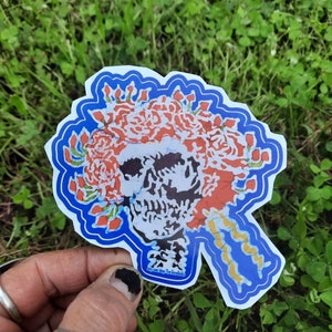 May include: A sticker featuring a skull with a wreath of red and white flowers. The skull is black and white, and the wreath is outlined in blue. The sticker is on a white background.
