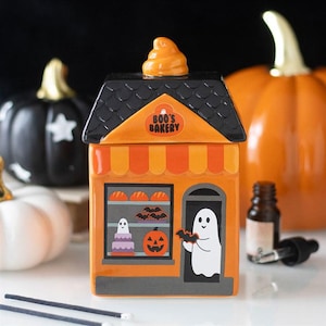 Peut inclure: Un pot en céramique en forme de boulangerie avec une façade orange, un toit noir et un couvercle surmonté d'un tourbillon orange. L'avant présente une enseigne BOO'S BAKERY, une vitrine avec des gâteaux, des chauves-souris, une citrouille et un fantôme.