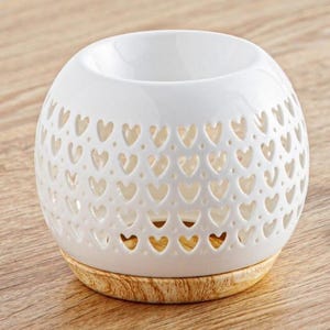 Op de afbeelding: Een witte keramische etherische olie diffuser met een ronde vorm en hartvormige uitsparingen. De diffuser staat op een houten voet. De bovenkant heeft een grote opening voor het toevoegen van olie en water.