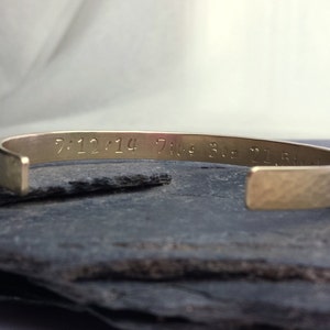 May include: A gold-toned metal cuff bracelet with a hammered finish. The bracelet has an inscription that reads "7.12.14 7:00 3.0z 21.5"