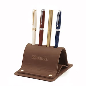 Puede incluir: Un portalápices de cuero marrón con cuatro bolígrafos. Los bolígrafos son blancos, rojos, dorados y azules. El portalápices tiene la palabra "Zhanestha" grabada en relieve en la parte delantera.