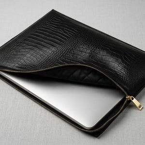 Puede incluir: Funda negra para portátil con un patrón texturizado similar a la piel de cocodrilo. La funda tiene una cremallera dorada y un tirador rectangular. Un portátil plateado es parcialmente visible.