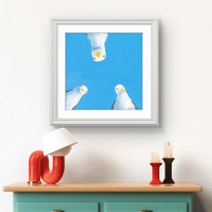 Könnte beinhalten: Ein gerahmter Kunstdruck mit drei Möwen vor einem leuchtend blauen Hintergrund. Die Möwen sind in Weiß und Grau mit gelben Schnäbeln dargestellt. Das Kunstwerk wird in einem weißen Rahmen präsentiert und verleiht dem Raum eine klare, moderne Ästhetik.