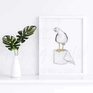 May include: A framed print featuring a watercolor illustration of a seagull standing on a roll of toilet paper. The seagull is white and gray with yellow feet. The print is displayed on a white shelf with a white vase and green leaves.