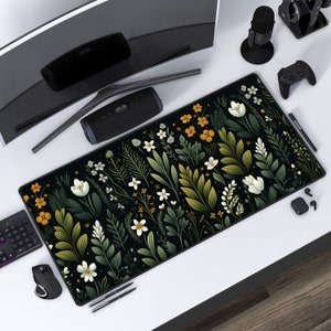 Op de afbeelding: Een zwarte en groene muismat met een bloemenpatroon, witte bloemen en gele accenten. De muismat ligt op een wit oppervlak met een computermonitor, een luidspreker, een gamecontroller en andere accessoires.